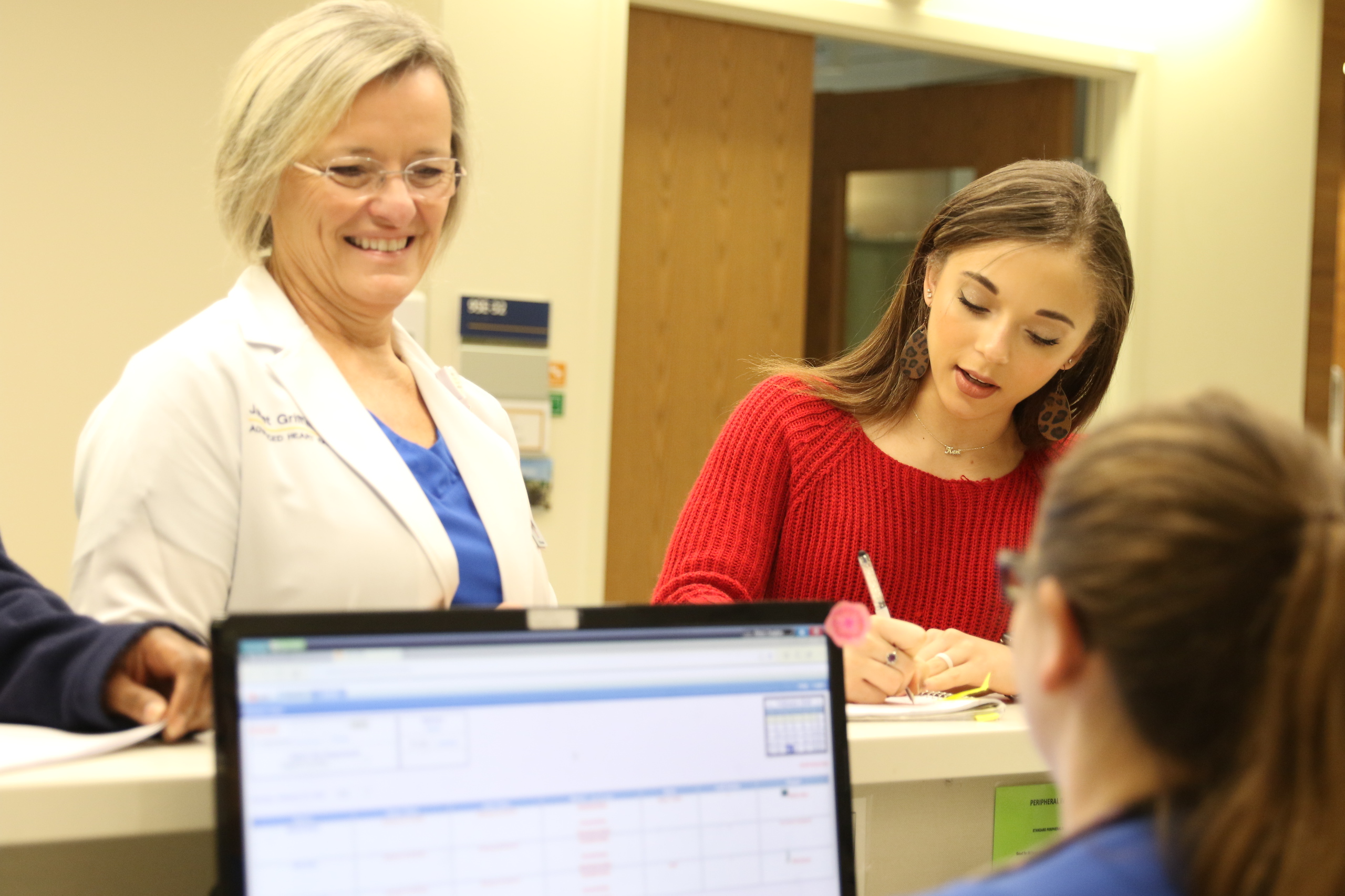 Female student with long brown hair in red shirt is standing at counter writing notes while medical provider stands beside her talking with person sitting at a computer behind the counter. 